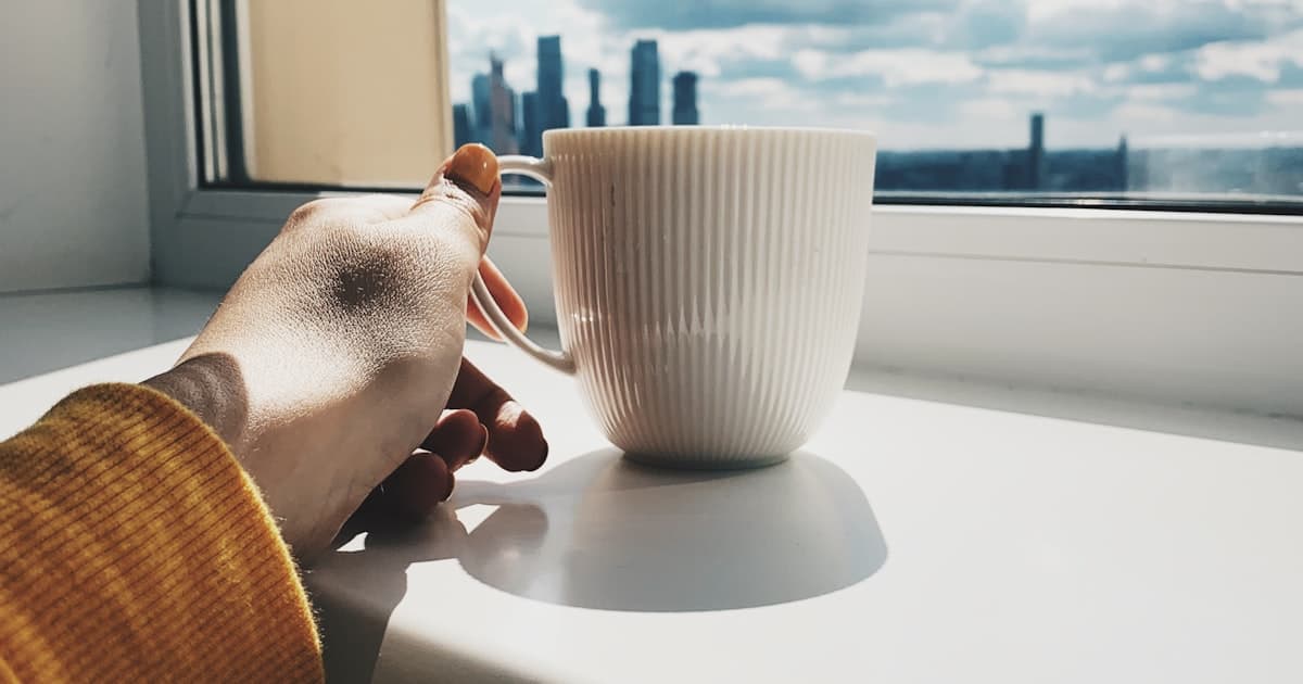 Une main attrape une tasse de café au soleil, près d'une fenêtre